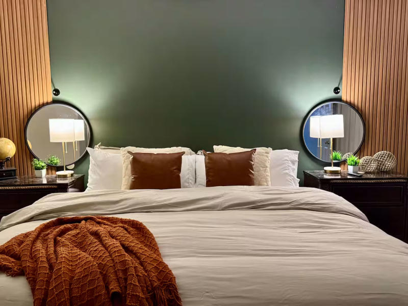 A stylish bedroom at Lakeside Haven with a modern design featuring a large bed with white linens, brown leather accent pillows, and a rust-colored knit blanket draped across the foot. The dark green accent wall behind the bed is framed with warm wood paneling. Two matching bedside tables hold lamps with white shades, round mirrors, small green plants, and decorative objects, creating a cozy and contemporary atmosphere.
