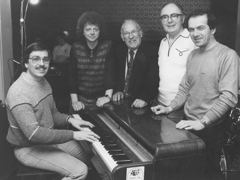 A black and white photo of five people around a piano in the 1990s.