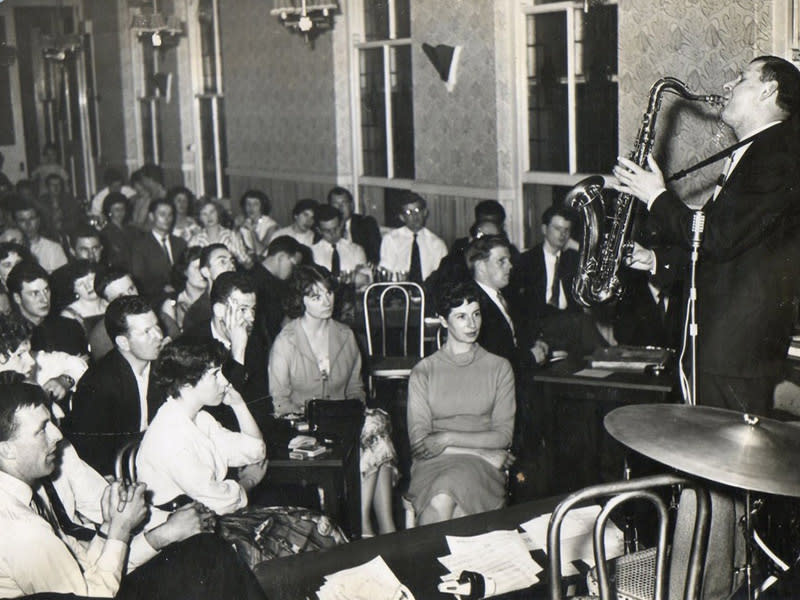 A black and white photo of a man playing saxophone in front of an audience.