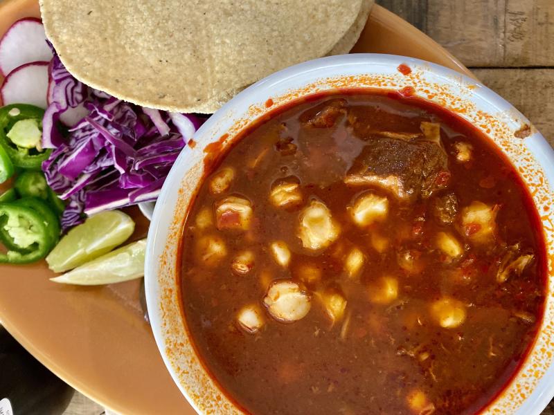 Close-up of a bowl of crimson red pozole rojo with hominy and tender pork in rich broth, served with a side of lime wedges, sliced radishes, shredded purple cabbage, jalapeños, and tostadas at Alta Baja Market in Santa Ana, California.