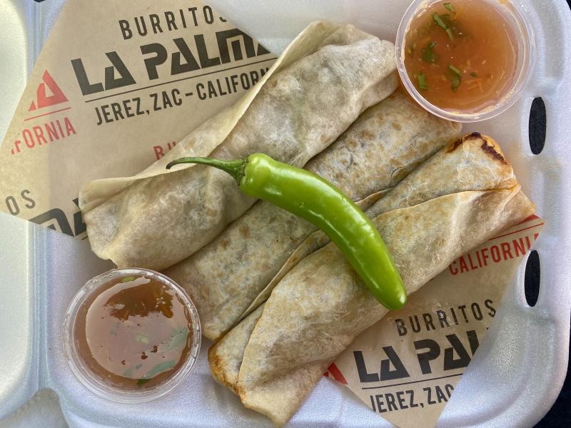Three slender Zacatecan-style burritos wrapped in soft flour tortillas, served in a red basket lined with Burritos La Palma-branded paper, accompanied by a fresh green chile pepper and two small cups of red salsa.