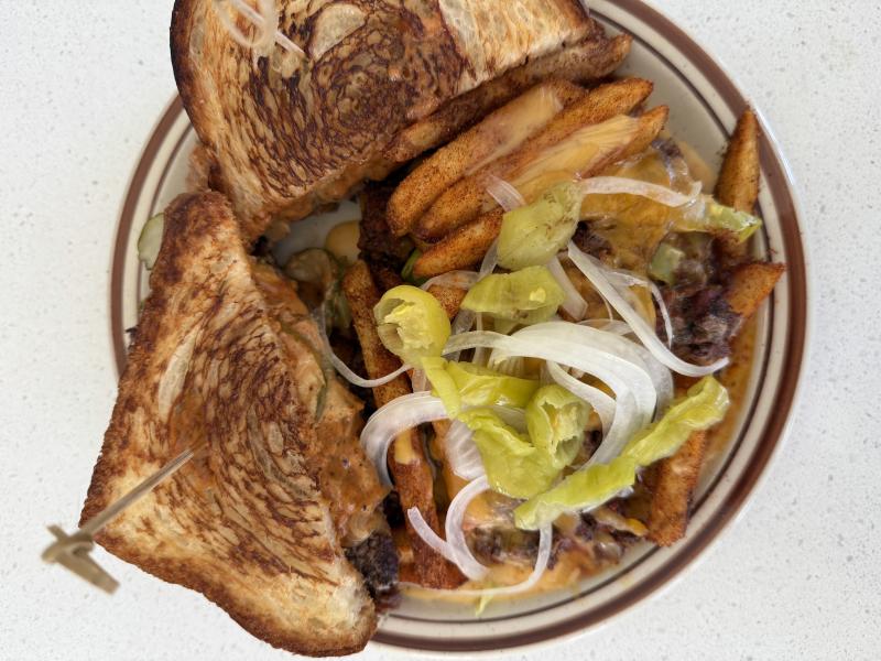 Grilled patty melt served with seasoned fries topped with melted cheese, sliced onions, and pickled peppers at Le Hut Dinette in Santa Ana, California.