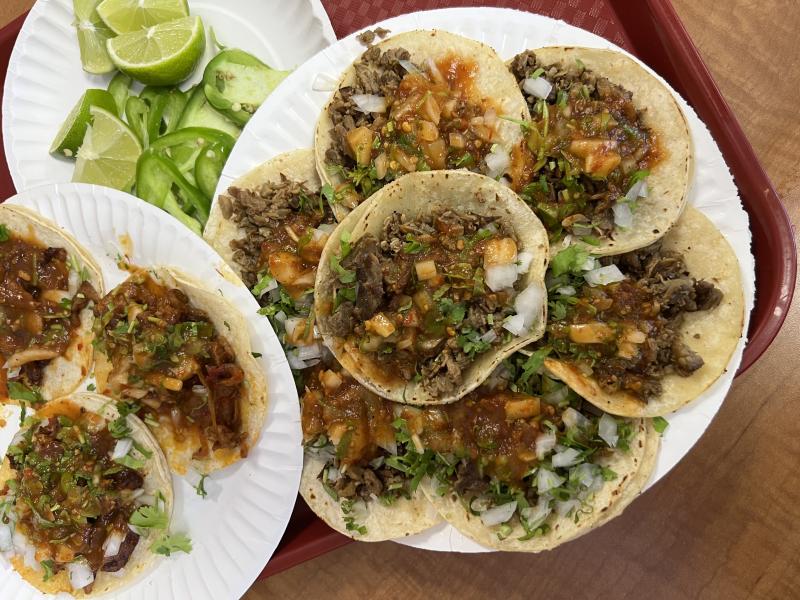 Plates of carne asada tacos topped with chopped onions, cilantro, and red salsa, served on corn tortillas with a side of lime wedges and sliced jalapeños on a red cafeteria tray at Taqueria De Anda in Santa Ana, California.