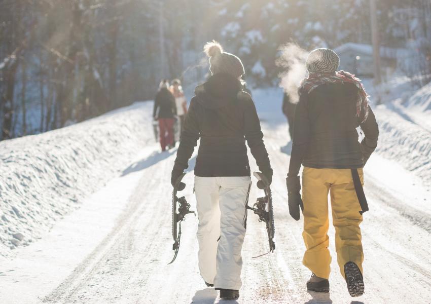 Two people carry snowshoes along a snowy, tree line path to a snowshoe trail