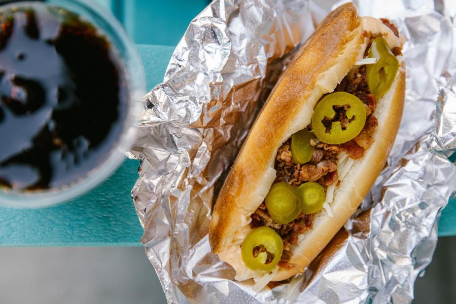A close-up of a foil-wrapped sandwich filled with meat and jalapeños, alongside a glass of dark soda on a teal table.