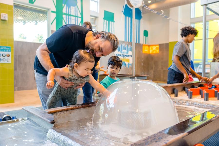 A family is at the Amazeum. A man is holding a baby, while a young boy looks on. The exhibit appears to involve water and interactive elements.