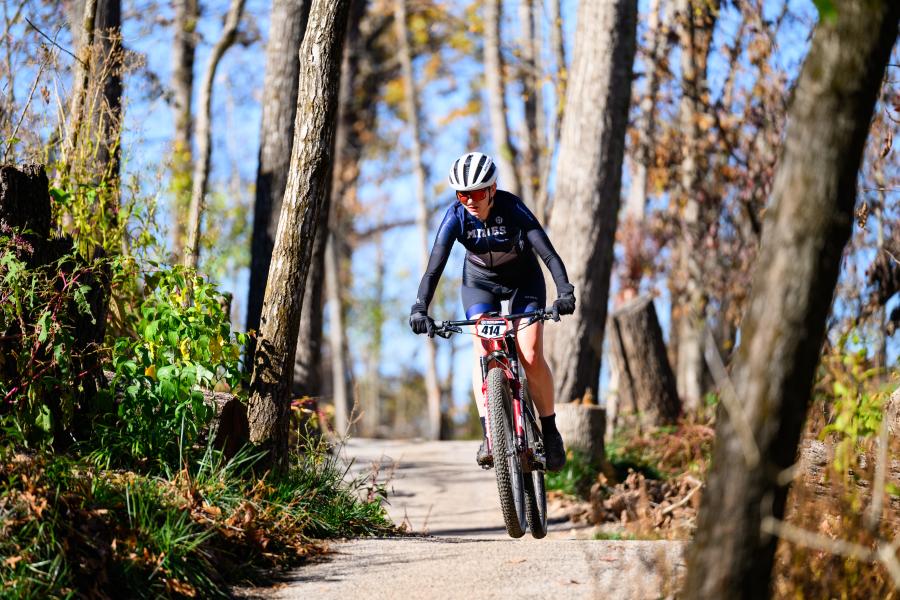 A woman in cycling gear rides a mountain bike on a dirt trail through a sun-dappled forest. She wears a helmet and sunglasses.