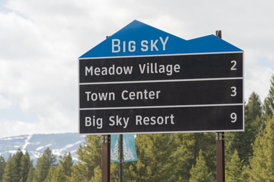 Signpost for Big Sky, listing distances to Meadow Village (2 miles), Town Center (3 miles), and Big Sky Resort (9 miles), surrounded by trees and mountains.