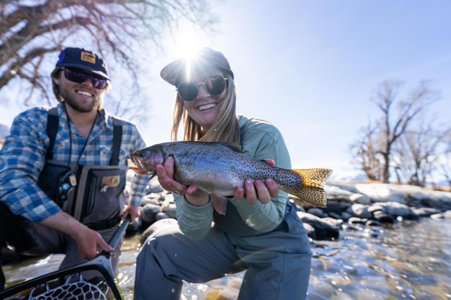 A woman holds a rainbow trout while smiling, with a man in the background. They are by a river, surrounded by trees and rocks.