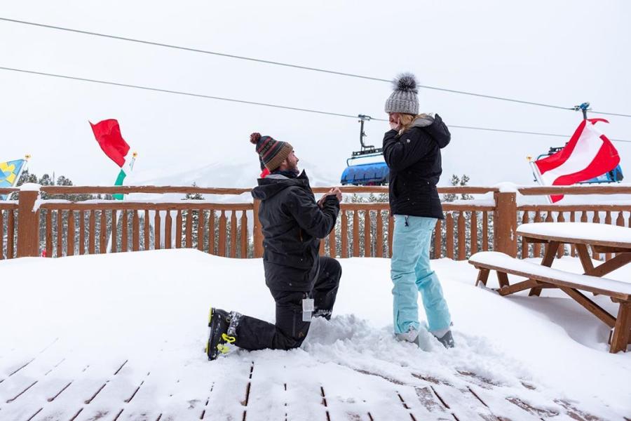 A person kneels in the snow, proposing to another on a wooden deck with ski lifts in the background and flags fluttering. Snowy mountains loom in the distance.