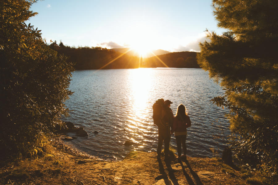 The sun is shining through a mountainous landscape onto a shimmering lake with two people standing by the lakeshore looking out.