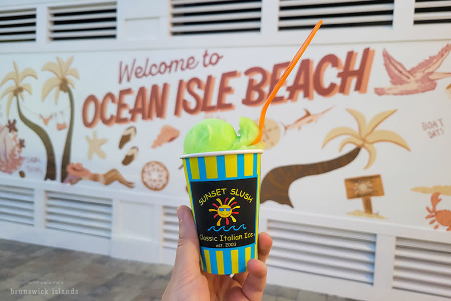 a cup of green italian ice from Sunset Slush pictured in front of a mural on Ocean Isle Beach