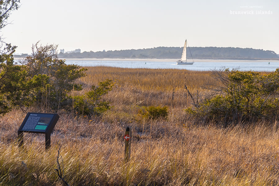 a view of a trail along marsh with an informational sign, directional sign, and a sailboat floating past in the background