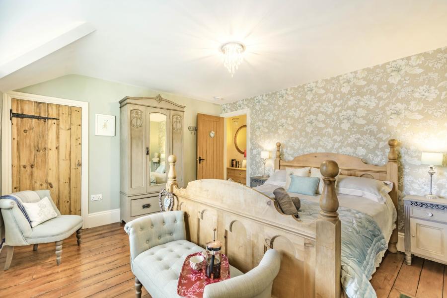 Bright country-style bedroom with a wooden bed, floral wallpaper, pale wood floors, and soft blue accents, creating a cozy, inviting feel.