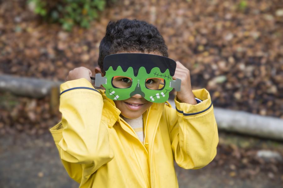 Child wearing a bright yellow raincoat and green monster mask at Snowshill Manor in the Cotswolds during a Halloween-themed activity.