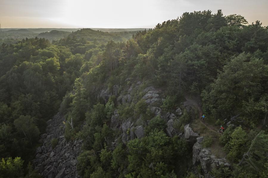 An aerial view of a couple of bikers on the RAMBA trail system. The trail features rock outcroppings and is surrounded by lush green foliage