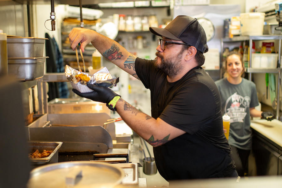bearded man in ball cap with tongs getting food out of a big pot on stove