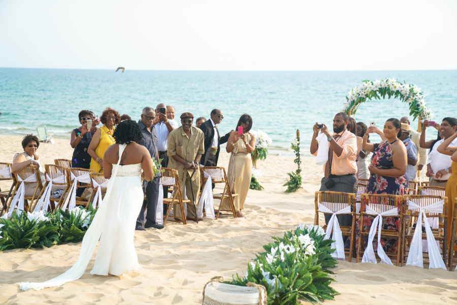 Bride walks down sandy beach aisle toward guests with sea view. Guests, dressed in bright attire, take photos. Floral arch and chairs enhance the festive atmosphere.
