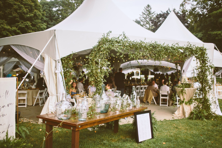 Outdoor wedding reception under white tents adorned with greenery. A wooden table with glass vases is in the foreground. Guests sit at tables, creating a festive atmosphere.
