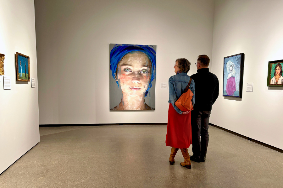 A couple views paintings in a gallery. The centerpiece is a large portrait of a woman with a blue headscarf. The room is bright and minimalist.