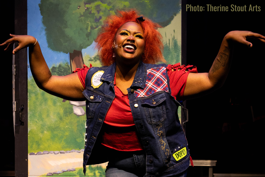 woman with orange hair acts out character on stage wearing red shirt, jean vest with colorful patches