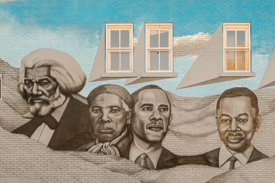A mural on a brick wall depicts four historical figures, Frederick Douglass, Harriet Tubman, Barack Obama and local muskegon leader Charles Waugh, in gray tones against a sky-blue backdrop. Two windows with triangular shadows are above them.depicting