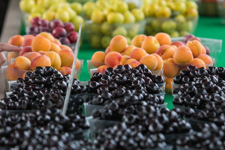 A market stall displays an array of fresh fruits, including dark cherries in the foreground, ripe peaches in the middle, and green grapes in the background. The fruits are neatly organized in clear containers on a green tablecloth.