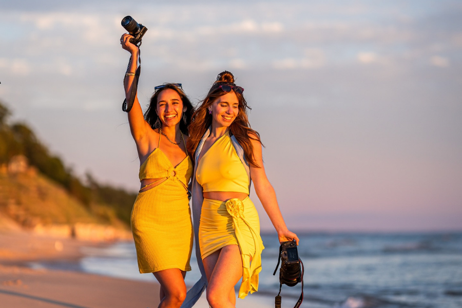 Two people walking on the beach at sunset, one holding a camera, both smiling and dressed in yellow outfits.