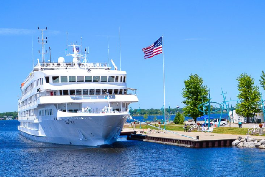 A large white cruise ship docked at a sunny pier, a U.S. flag flutters to its right; behind are trees, cars, and a clear blue sky.