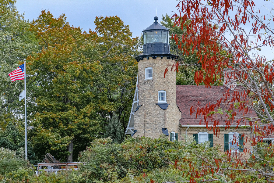 Historic stone lighthouse with black lantern tower and red-roofed building, beside an American flag amid colorful autumn foliage.