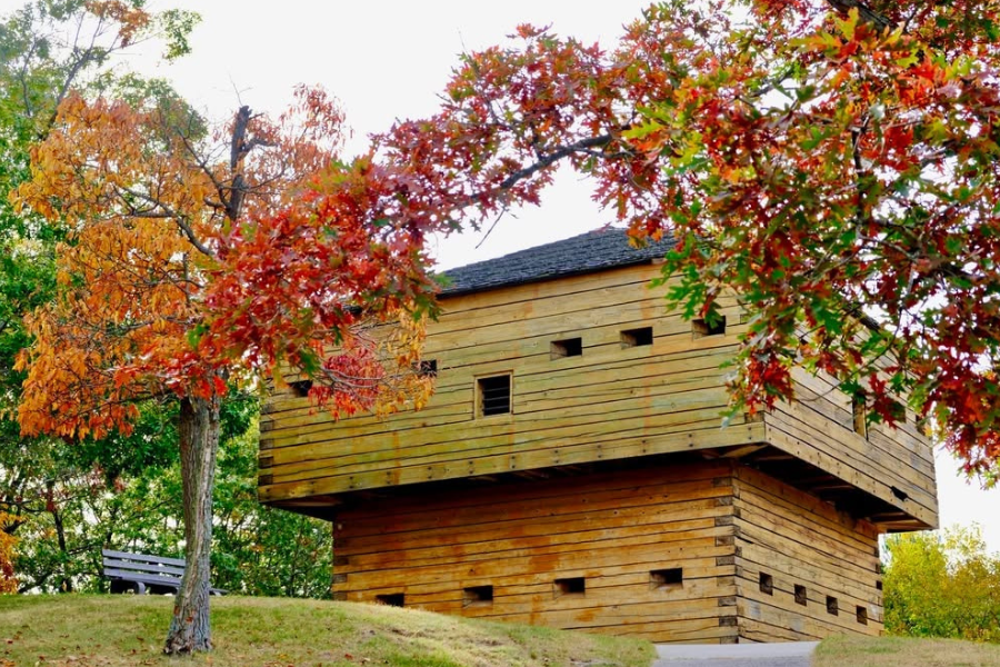 Historic wooden fort-style building with small square windows, framed by vivid red and orange autumn trees under a bright sky.