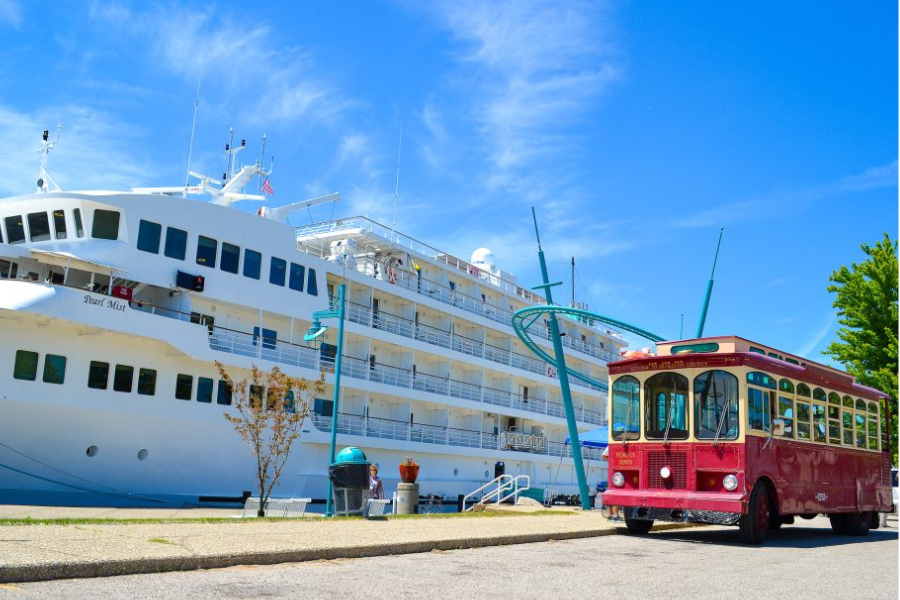 A red trolley bus parked beside a large white cruise ship under a bright blue sky, conveying a sense of travel and leisure on a sunny day.