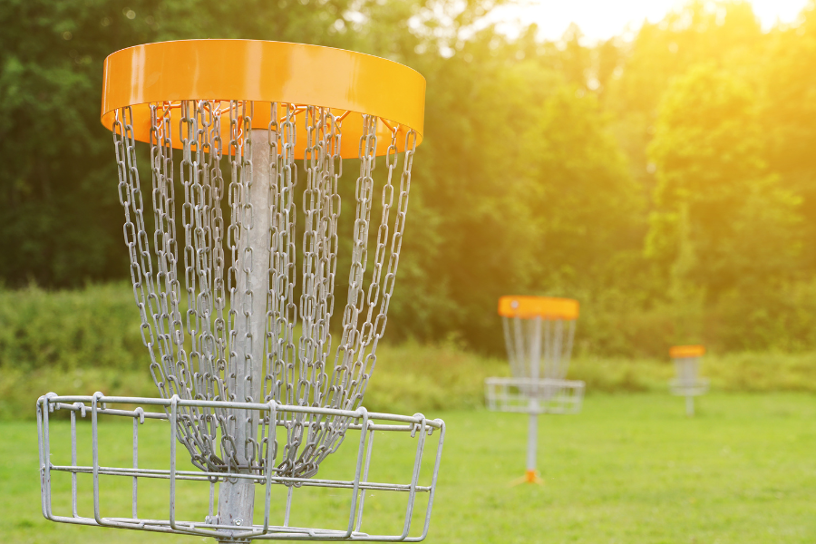 3 yellow rimmed disc golf baskets in green field are backlit by warm sunlight. green trees line the horizon