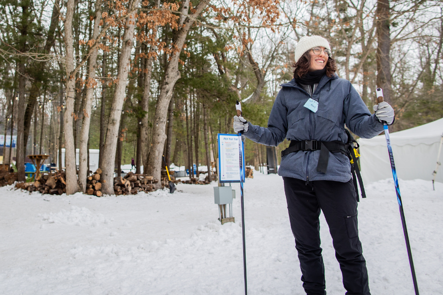 A person in winter gear and a white beanie smiles while holding ski poles in a snow-covered forest. Trees and a tent are visible in the background.