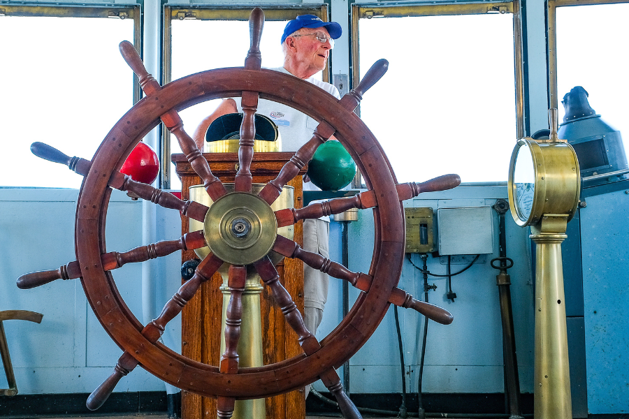 man in blue baseball cap stands behind wooden captains wheel in brigde of milwaukee clipper museum ship