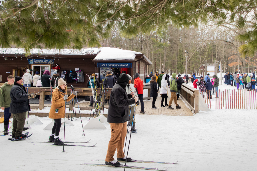 People in winter clothing rent and use ski equipment near a snow-covered rental hut. Trees and a light-hearted crowd create a lively, outdoor atmosphere.
