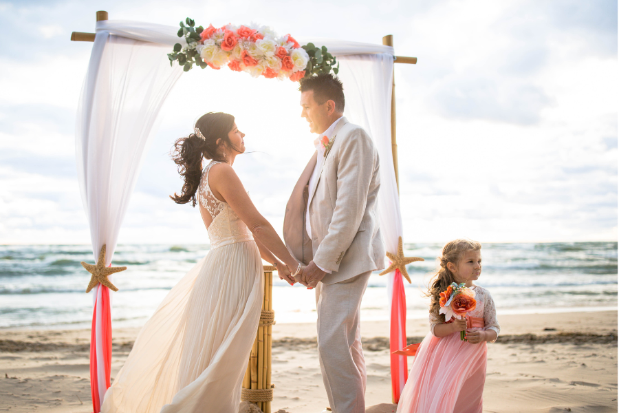 A bride and groom hold hands under a floral arch on a beach, dressed in white and beige. A young girl in pink stands beside them, holding flowers.