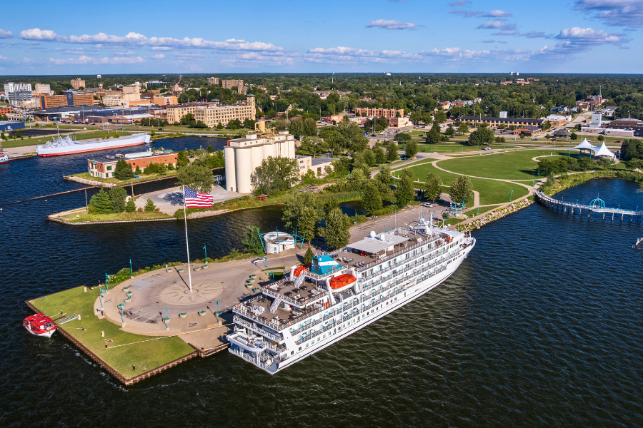 Aerial view of a large white cruise ship docked at a vibrant lakeside city. A giant American flag waves, surrounded by green parks under a blue sky.