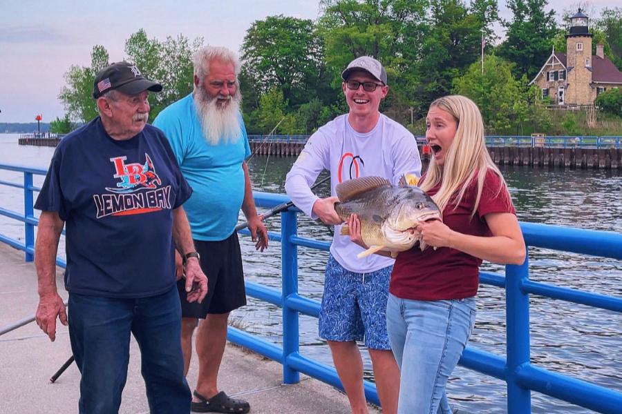 Four people on a pier, three observing the fourth person who is excitedly holding a large fish. In the background is a lighthouse.