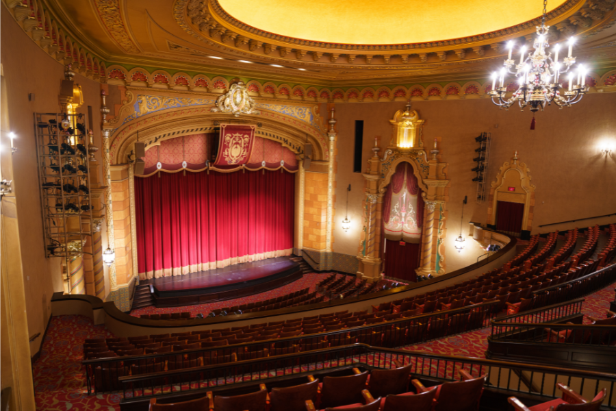 Interior view of historic theater from balcony, overlooking seating and elaborately ornamented stage with red curtains.