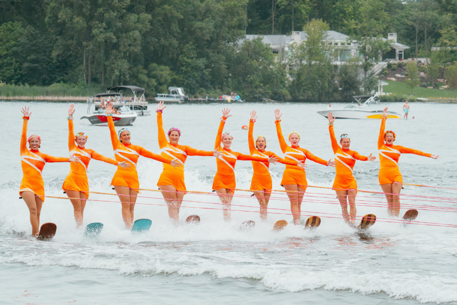 A group of water skiers in orange uniforms performing a line formation on a lake, with boats and trees visible in the background.