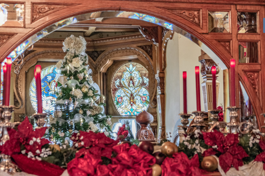 Elegant holiday setting with a decorated Christmas tree and ornate stained glass reflected in a carved wooden mirror, surrounded by red floral arrangements and candlesticks.