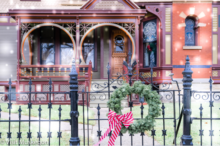 An ornate Victorian house in Muskegon with detailed woodworking, stained glass windows, and a prominent porch framed by an intricate metal fence adorned with a green wreath featuring a red bow.