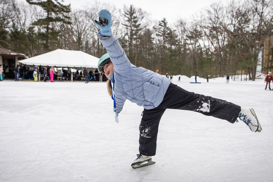 A person in a light blue coat and black pants balances on one ice skate, arm outstretched, smiling. Snowy trees and a tent with people are in the background.