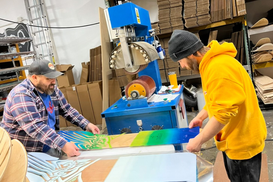 Two individuals working with a printing press in a workshop, collaborating on producing a colorful Snurfer snowboard.