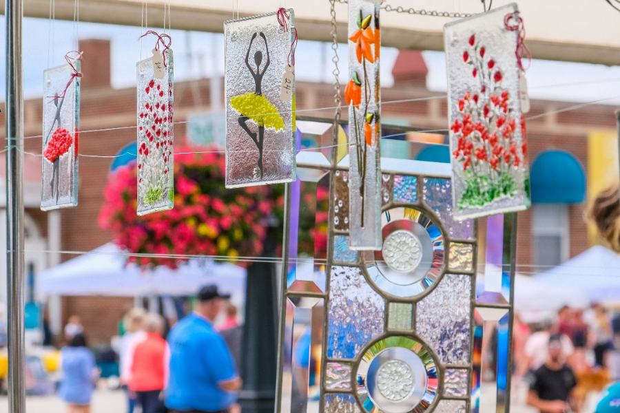 A variety of colorful glass art pieces and wind chimes hanging at a bustling Lakeshore Art Festival.