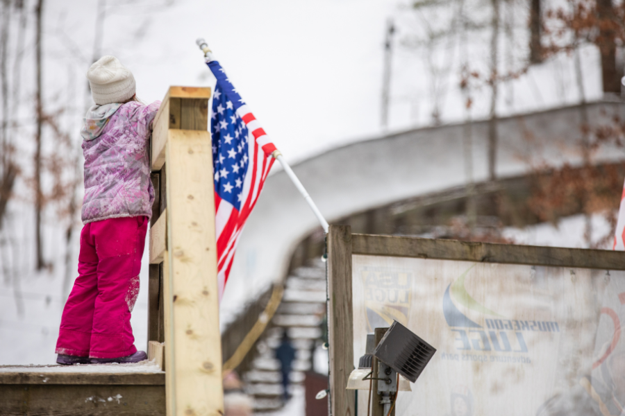 A child in pink winter clothes stands on a snowy platform near an American flag. In the background, a curving luge track extends into snowy woods.