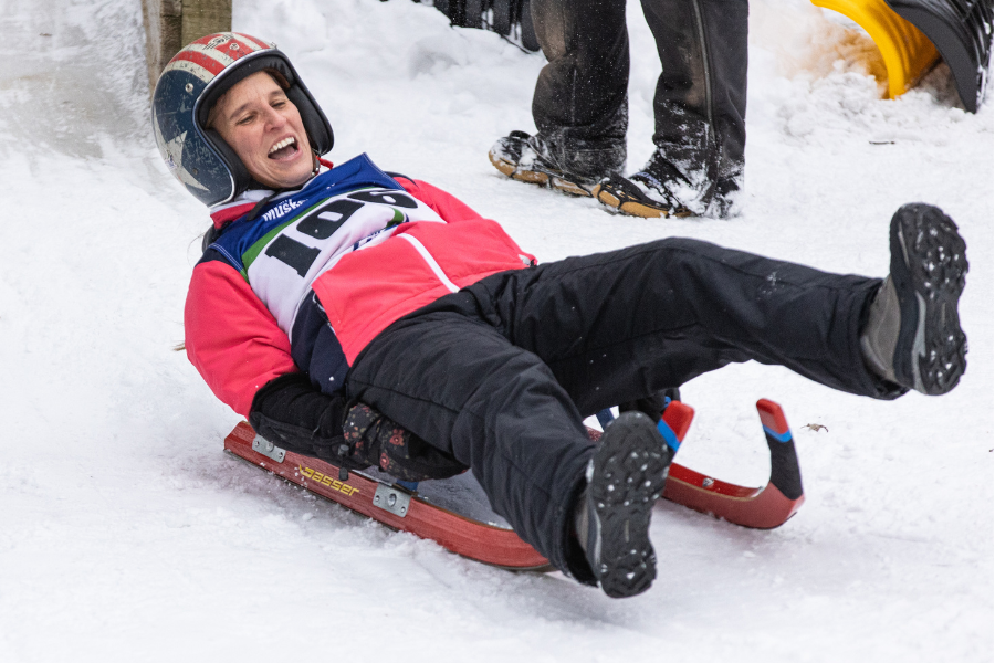 A person on a luge sled enjoys a ride down a snowy track, smiling widely. They wear a helmet, a red jacket, and black pants. The scene conveys excitement and joy.