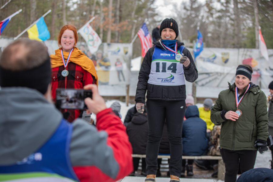 Three athletes stand on a podium in winter attire, smiling and wearing medals. A person in a red jacket takes a photo. Flags and trees in the background.