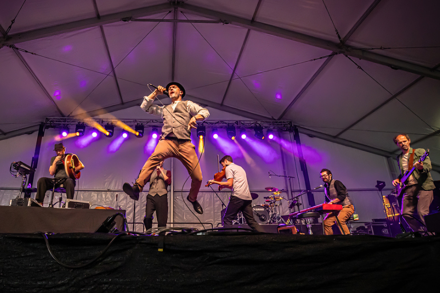 Person energetically performing on stage with a microphone at a live concert, accompanied by four musicians playing guitar, violin, and keyboards under a well-lit tent at Michigan Irish Music Festival